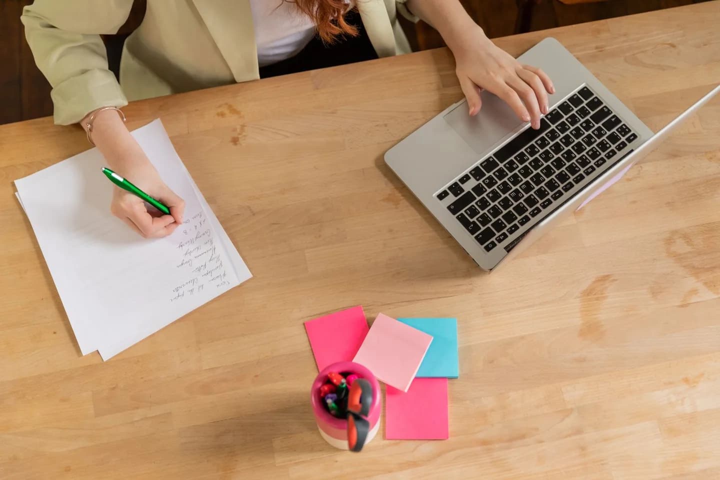 A person writing notes beside a laptop on a wooden desk