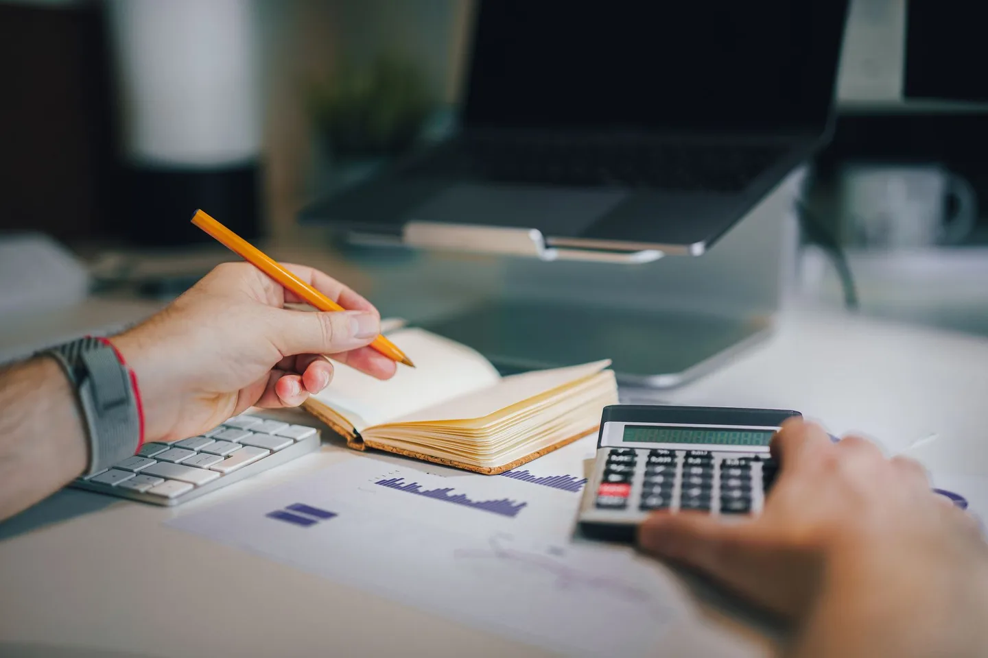 Hands taking notes and using a calculator beside a laptop